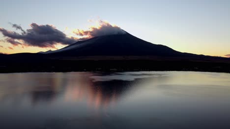 Dawn-breaking-over-a-tranquil-lake-with-a-majestic-mountain-backdrop,-clouds-caressing-the-peak