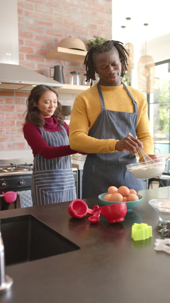 Vertical video of happy diverse couple in aprons baking in kitchen at home, slow motion