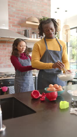 vertical video of happy diverse couple in aprons baking in kitchen at home, slow motion