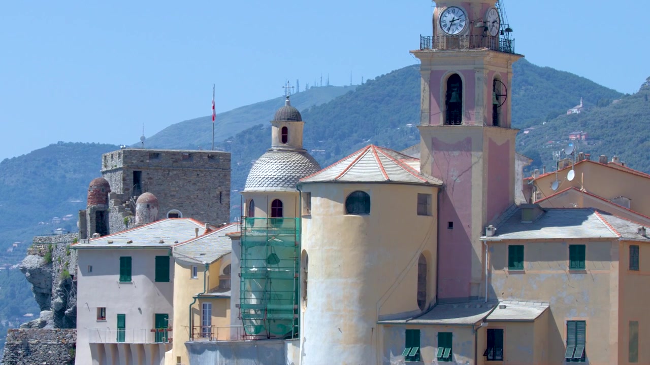 Una lenta inclinazione verso l'alto rivela edifici mediterranei colorati e una torre campanaria storica sotto la luce intensa del sole