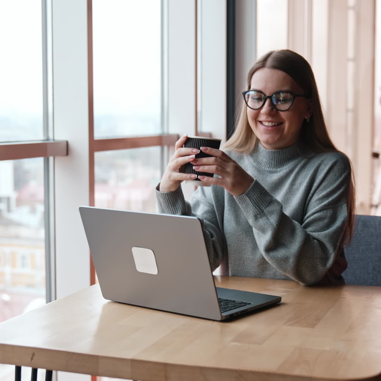 Cheerful lady in glasses having conversation via internet. Woman with coffee cup finishes conversation and looks at window