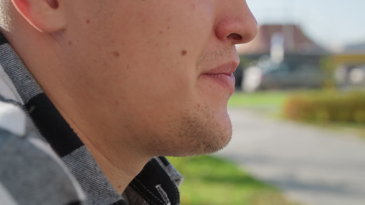 close up of white boy in checkered shirt exhaling calmly with gentle focus on lips and chin as sunlight touches skin in urban park setting with soft blur of road, greenery, and distant buildings