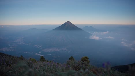 Agua-Volcano-towers-over-misty-valley,-sunset-view,-Antigua-below,-purple-flowers-in-the-foreground