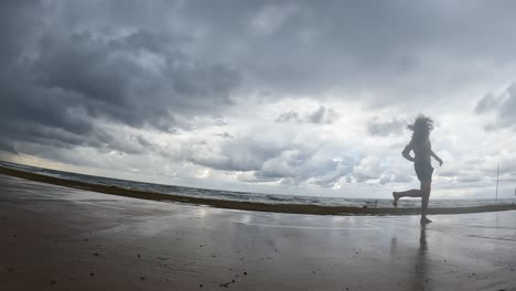 Zeitlupen-Mann-Mit-Langen,-Dicken,-Schwarzen-Haaren,-Der-An-Einem-Bewölkten-Tag-Am-Strand-Entlang-Läuft