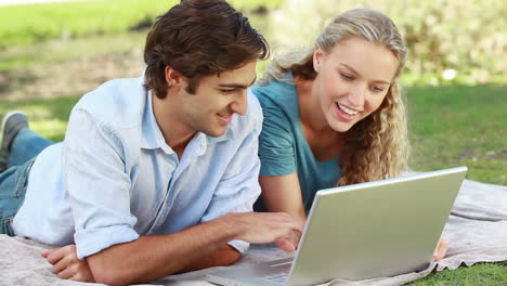 A-man-lies-with-a-laptop-as-his-girlfriend-lies-beside-him-and-they-look-into-the-camera