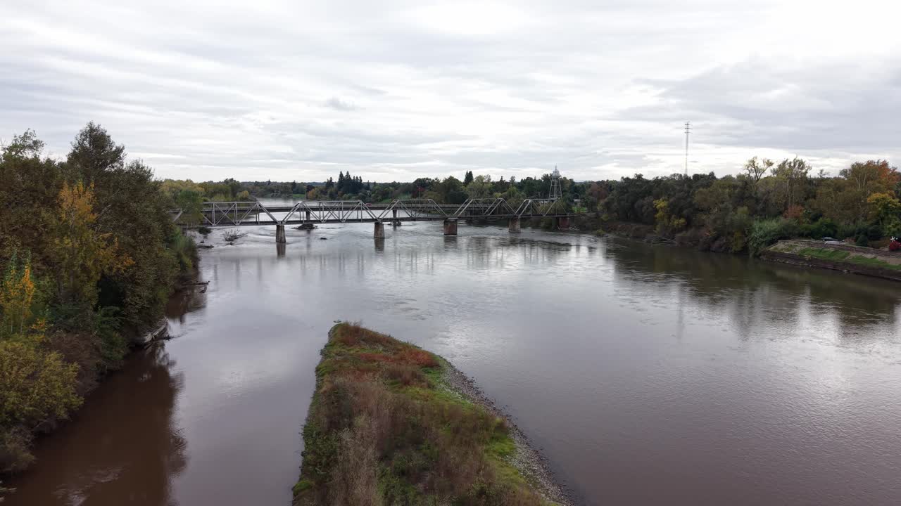 The drone glides above Mill Creek Park, revealing the Tehama Railroad Bridge stretching across the river with its historic steel frame highlighted by soft natural light