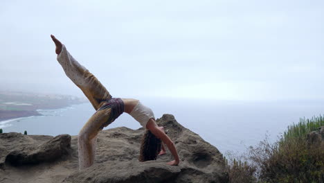 con el mar azul y el cielo azul como telón de fondo, una niña adopta una postura de puente mientras está de pie en una piedra, haciendo yoga en las rocas