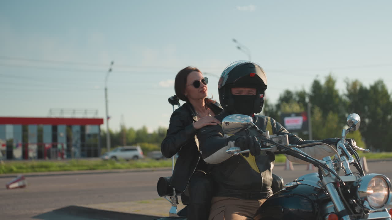 Motorcycle rider lifts helmet cover while female passenger in leather jacket steps onto bike, preparing to ride off as car passes by, bright sunlight flare highlighting motion