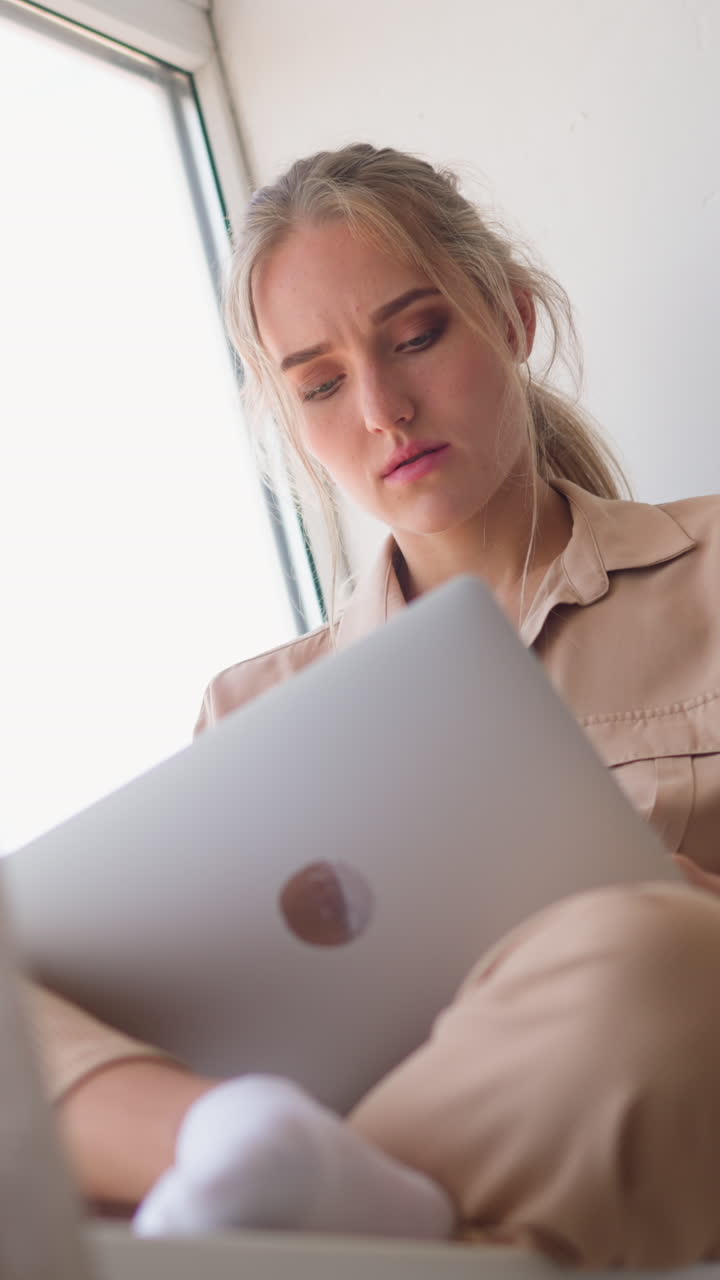 Young accountant woman makes calculations for work sitting on windowsill behind brown curtain leaned on pillow at home slow motion