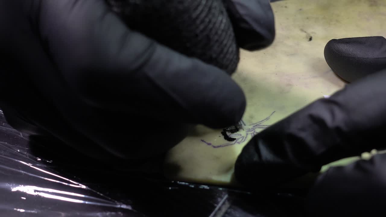 Inquadratura fissa di un tatuatore che lavora a un disegno su pelle sintetica, esercitandosi con un riferimento.