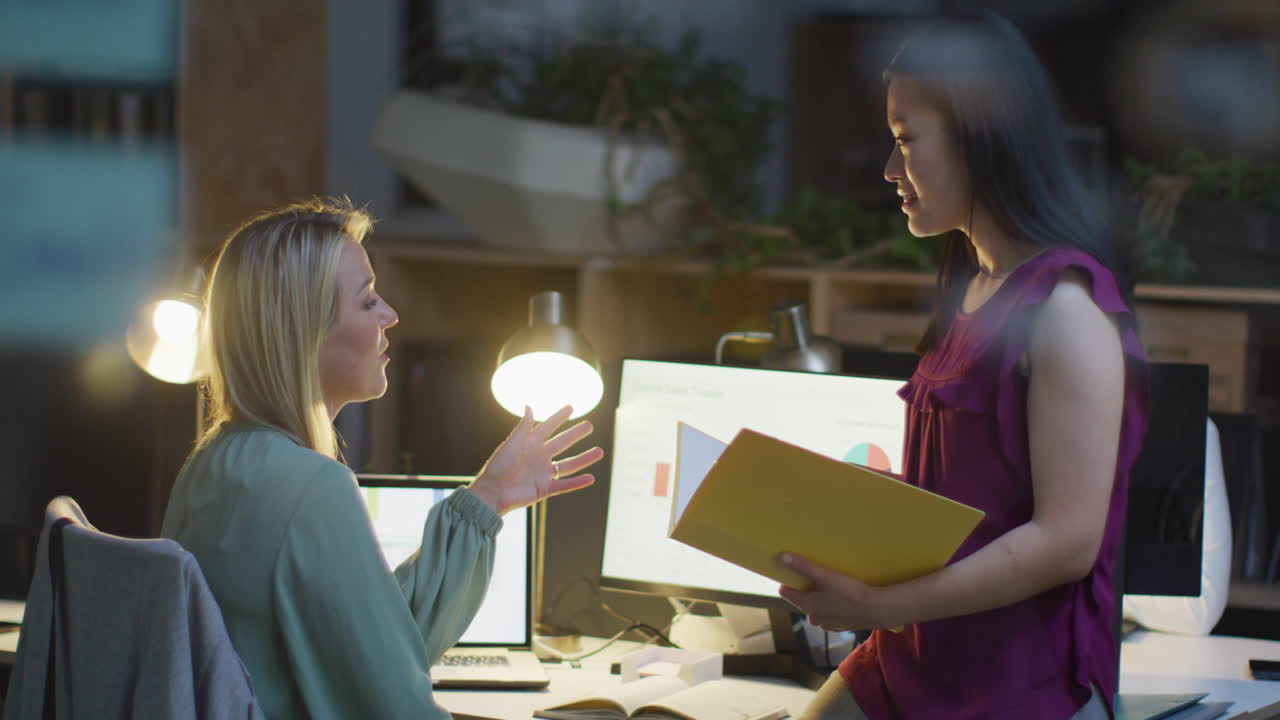 Caucasian and Asian businesswomen discuss work in an office working late