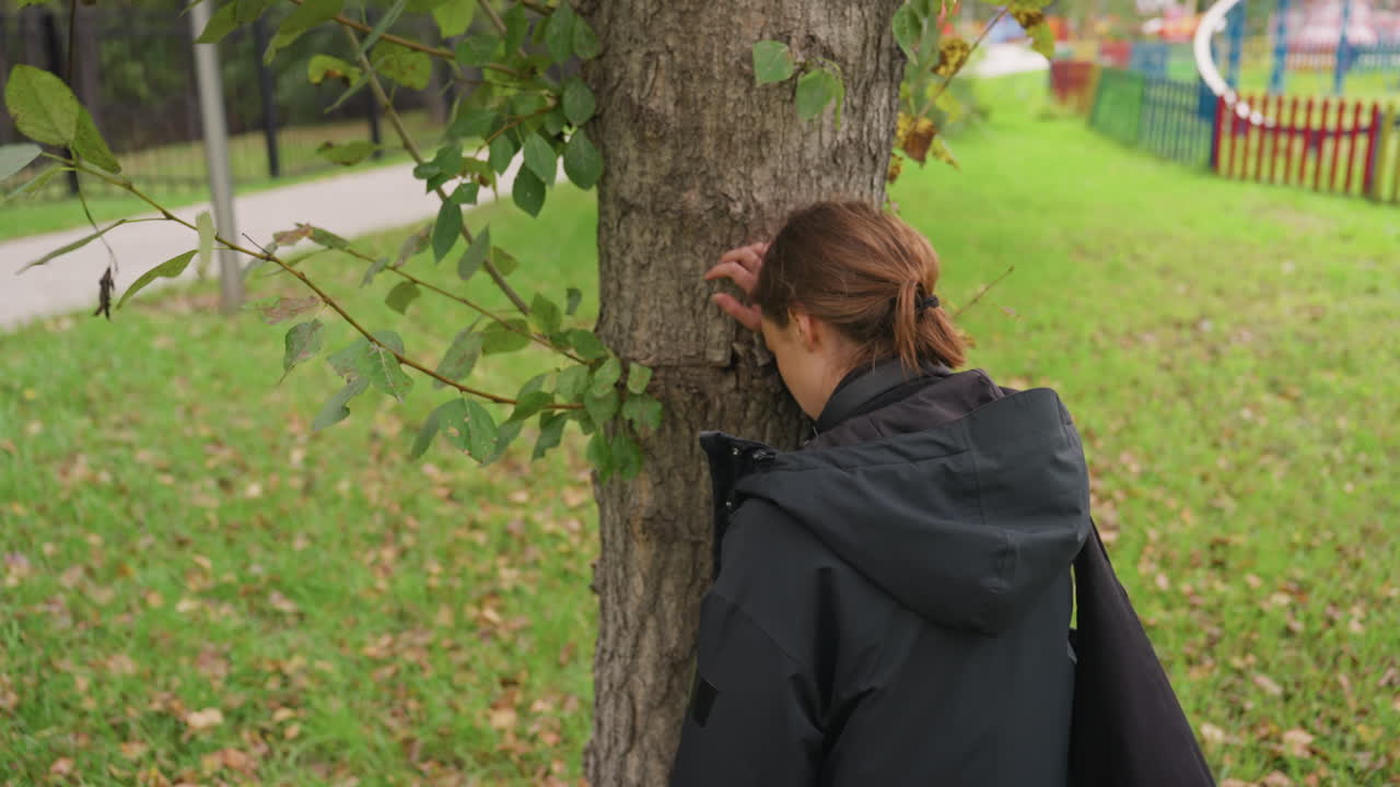 Woman leaning against tree trunk park, wearing dark rain jacket, hair tied back, head resting on bark, surrounded by fallen leaves on green grass, pathway and fence visible in background, overcast