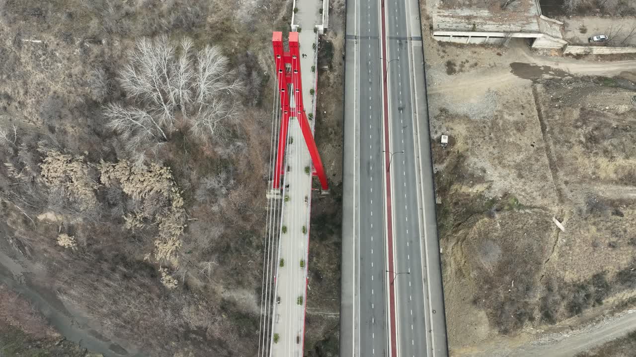 aerial view of a striking red bridge extending through a landscape of muted tones and winter trees. The design contrasts with the natural and urban surroundings