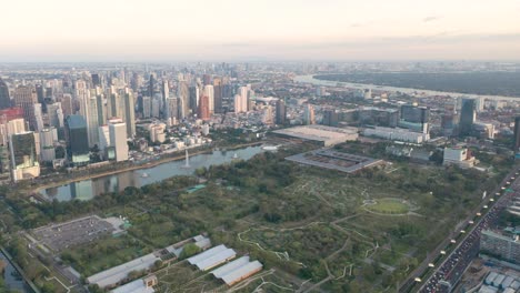 Aerial-drone-above-skyline-and-skyscrapers-of-Sukhumvit-district,-Pathum-Wan-and-Lumphini-Park-during-sunset-in-Bangkok,-Thailand