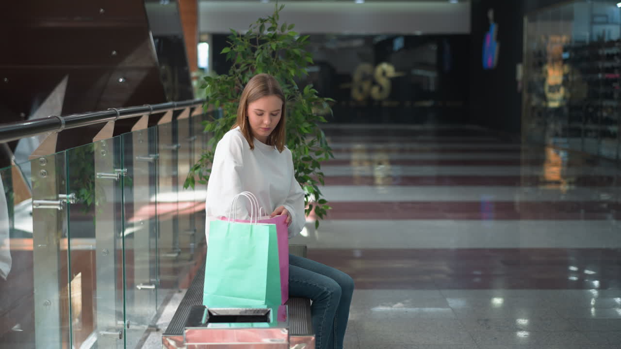 밝은 현대 쇼핑몰에 앉아있는 여성 쇼핑객은 핑크색 가방에서 노란색으로 인 책을 가져오고, 녹색 식물, 유리 레일링 및 넓은 반사 바닥으로 둘러싸여있는 동안 무작위로 열고 있습니다.