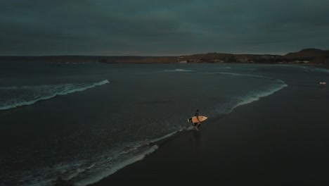 drone aerial of a man with a surfboard walking out of the ocean during sunset in peru