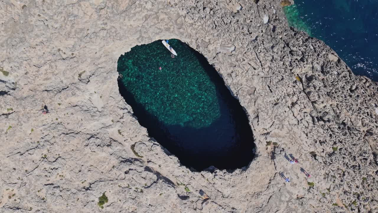 An aerial footage of a natural rock pool filled with clear turquoise water, surrounded by rugged limestone cliffs. People are swimming and kayaking inside the pool