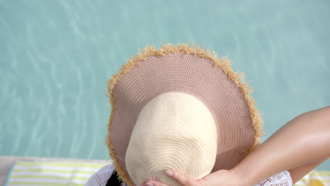 adolescente biracial con sombrero de sol sentada junto a una piscina soleada, en cámara lenta