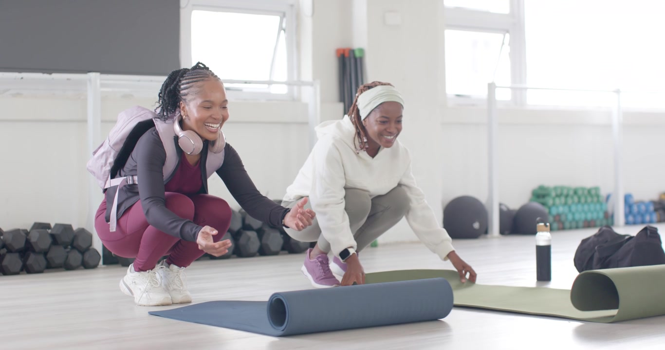 Two adult African women arriving at fitness studio unrolling blue, green mats, preparing to workout