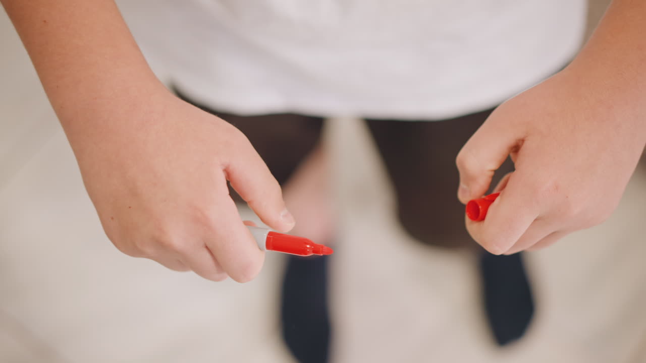 Tangan anak memegang tutup spidol merah, murid kecil melatih keterampilan motorik halus, jari-jari menggenggam dan memutar tutup, memeriksa ujung spidol, lantai ubin dan kaus kaki terlihat, momen konsentrasi yang tenang.