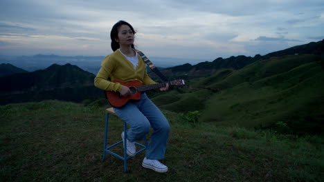 with the mountains in the distance, a woman sits atop a hill, her fingers expertly plucking the strings of her guitar as she plays a beautiful melody