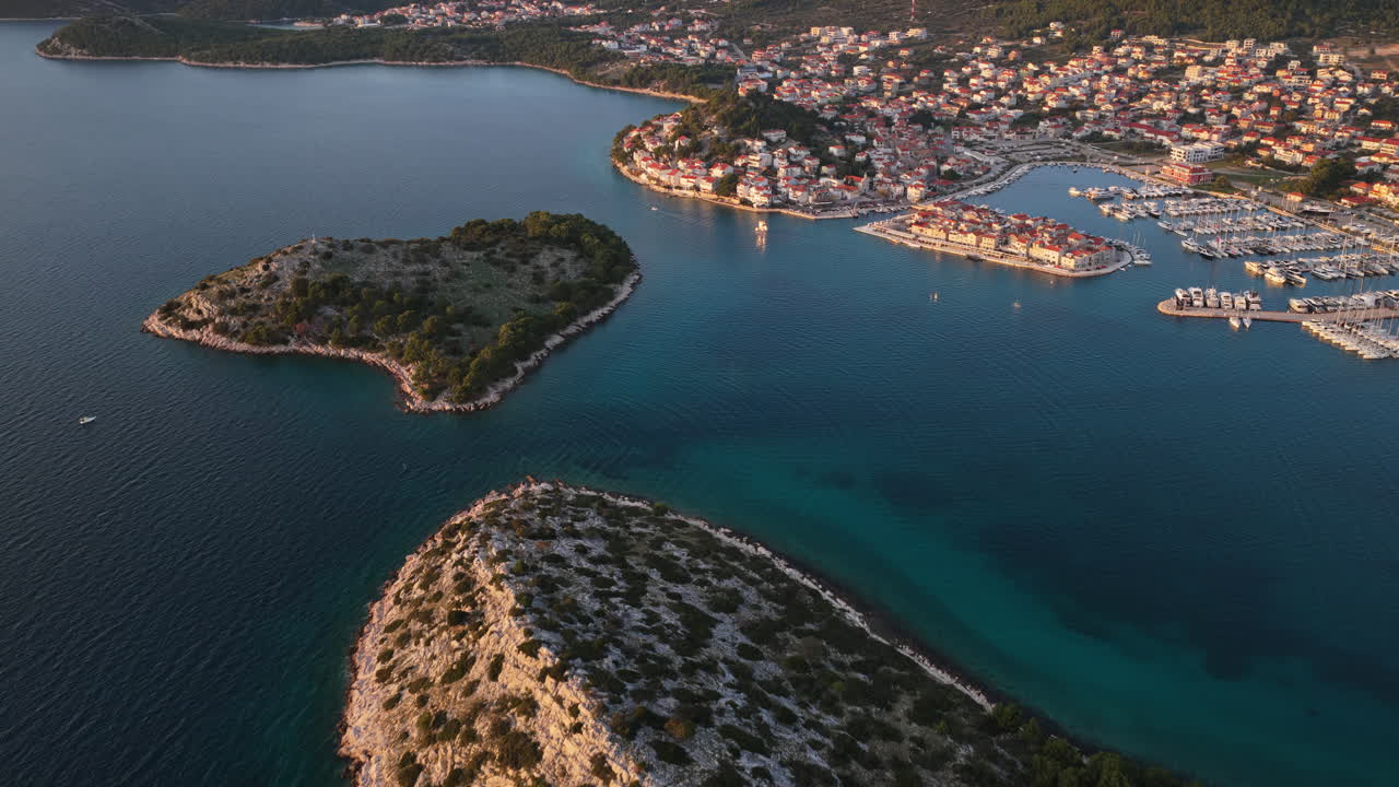 Aerial drone view of rocky green islands separated by narrow channels of turquoise water, with a coastal town and marina visible in the background. Tribunj, Croatia