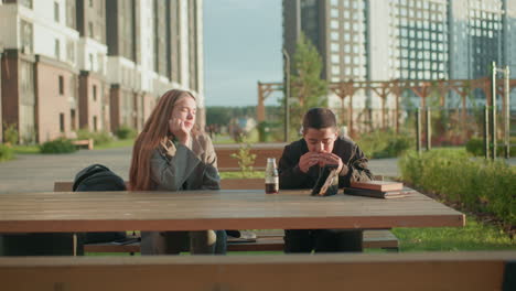Mum watching son enjoy shawarma while sitting together at outdoor wooden table in modern residential area, boy focused on meal with juice bottle and sandwich pack under warm sunlight