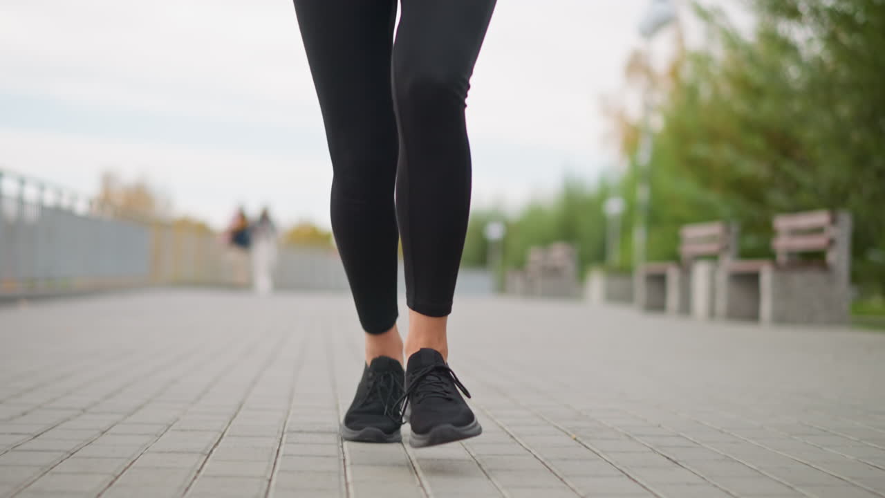 Close-up view of woman's legs in black leggings and sneakers running along park path, with blurred background of people walking, benches, and trees, promoting active lifestyle and fitness