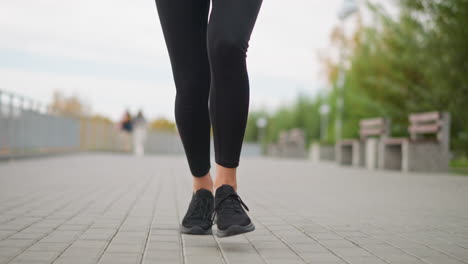 close-up view of woman's legs in black leggings and sneakers running along park path, with blurred background of people walking, benches, and trees, promoting active lifestyle and fitness