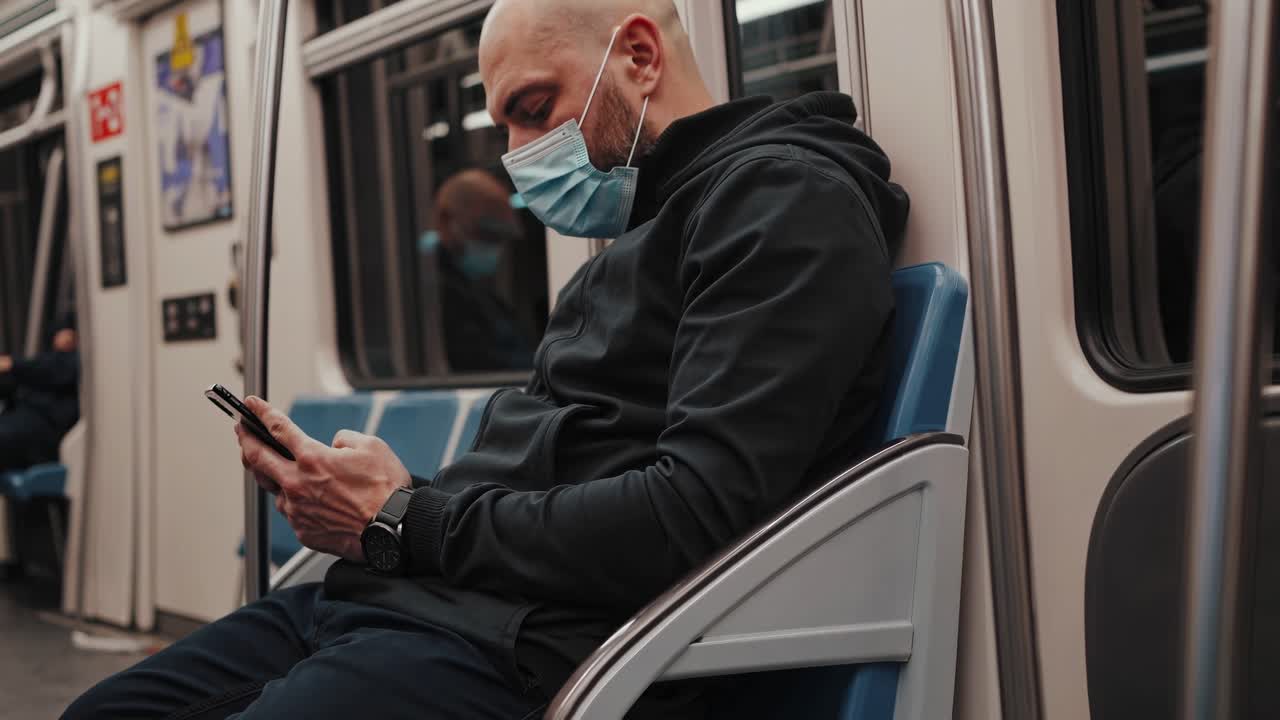 Commuter wearing protective face covering using smartphone while sitting inside subway train carriage during pandemic era, representing modern urban travel and communication