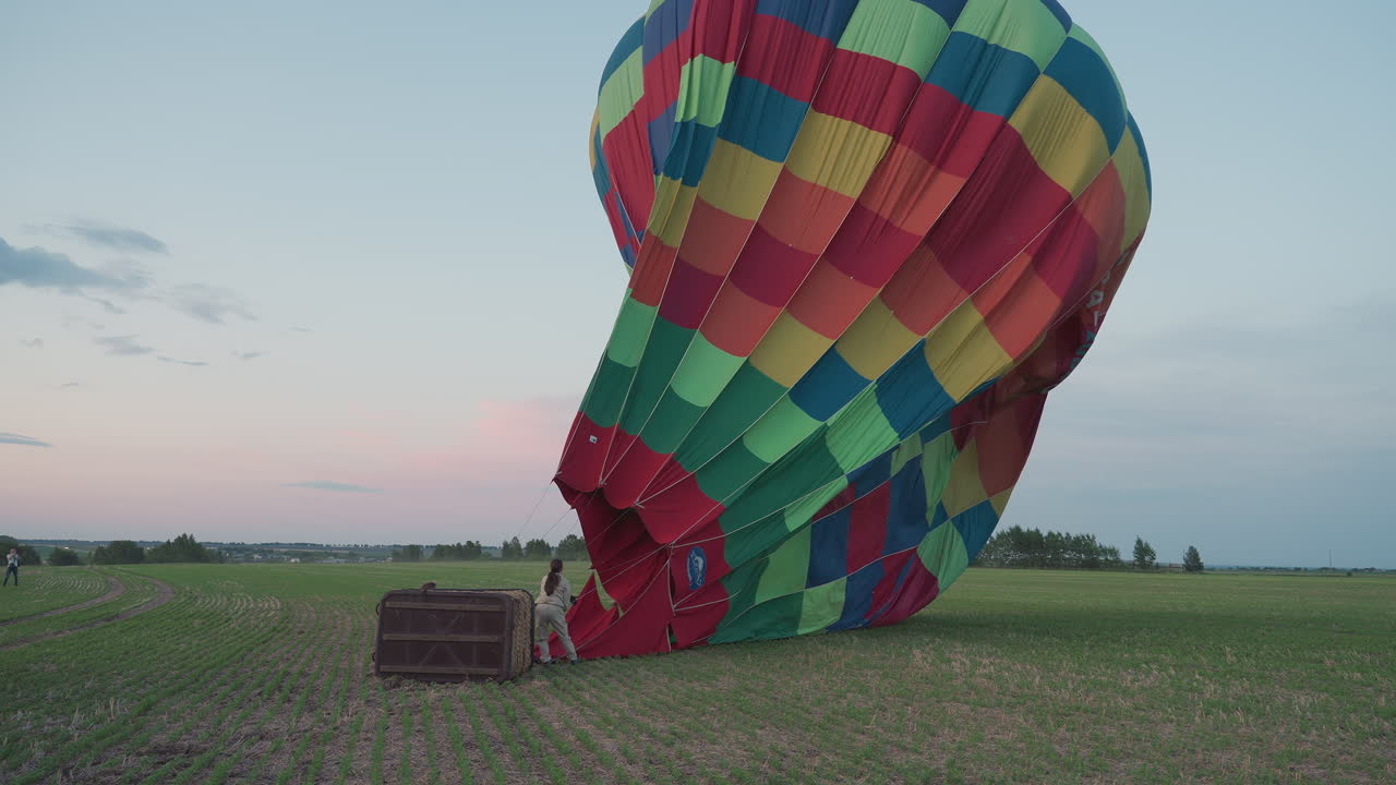woman pulling deflated colorful hot air balloon canopy across green farmland toward waiting trailer under soft dusk sky creating careful dynamic landing