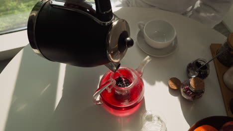 pouring hot water into teapot on table closeup. brewing hibiscus tea with flow from electric kettle for breakfast in shady kitchen. healthy beverage