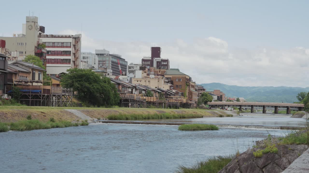 Scenic view of the Kamo River in Kyoto, Japan with peaceful flow of water, surrounded by traditional housing and lush nature under a bright sunny sky.