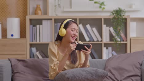 young women listening to music in headphones and dancing energetic and rhythm at couch. attractive asian girl looking at the smart phone sitting in the living room spending leisure time.