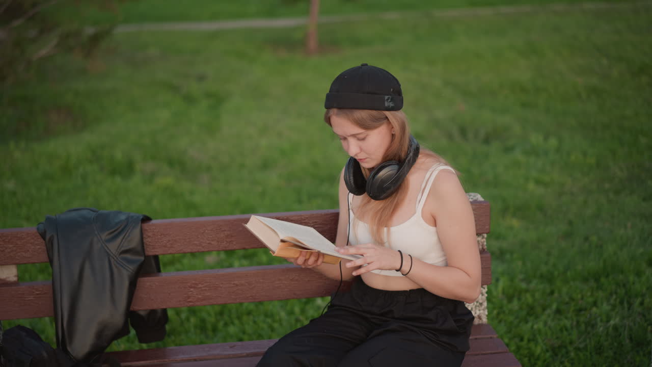 young white student reading book on wooden bench in green park, headphones around neck and jacket beside her, turning pages with concentration, soft daylight and casual street study vibe