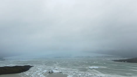 Vista-Sobre-La-Laguna-Glaciar-Islandesa-Con-Bloques-De-Hielo-Que-Fluyen-Y-Miradas-A-La-Montaña-De-Hielo-Y-Al-Cielo-Blanco-Brumoso