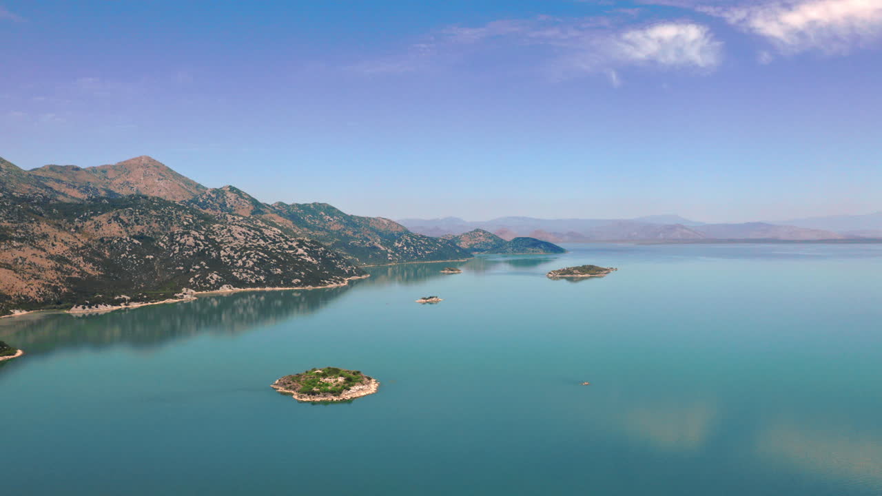 Breathtaking view on lake Skadar in Montenegro with its crystal clear blue water reflecting the sky, with tall mountains on the left side and a hazy horizon in the distance, scenic aerial shot 4K