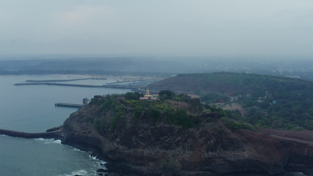 Drone pans right to left across Ratnadurga Fort and Shri Devi Bhagwati Temple on seaside cliff, revealing harbor breakwater, hazy ocean, waves, and coastal expanse