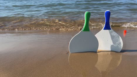 Beach-tennis-rackets-and-red-balls-on-sandy-beach-with-waves-breaking-on-shore