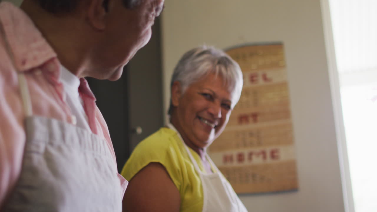 Felice coppia anziana birazziale che indossa i grembiuli, prepara il cibo e sorride.