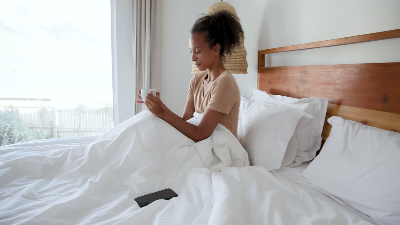 Donna che si rilassa a letto, gustando il caffè e navigando sullo smartphone al mattino, a casa