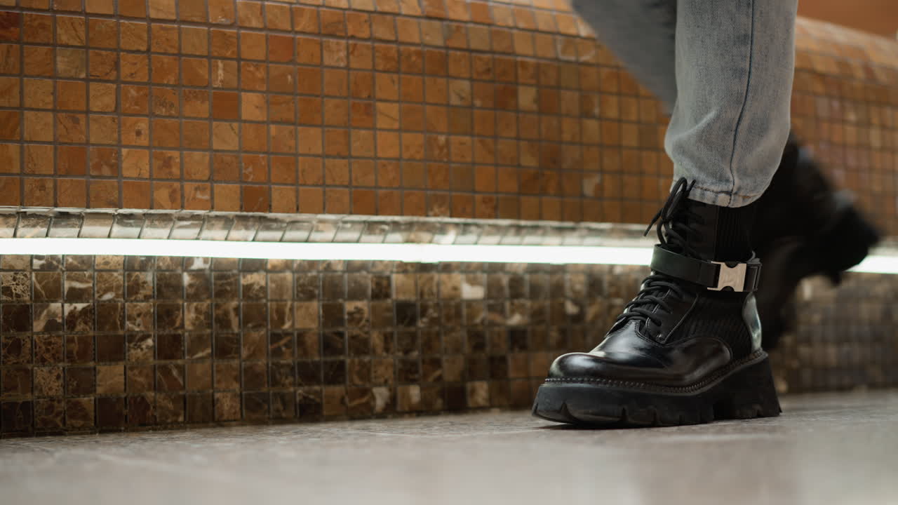 Low angle close up shot of shopper wearing black leather boots stepping onto mosaic bench seat near flowing mall fountain, showcasing stylish leg motion in modern indoor shopping center