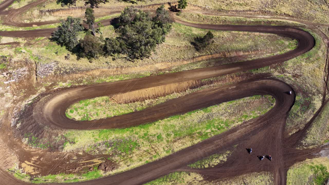 Multiple dirt bikes navigate sharp turns on a winding dirt track in rural Australia, captured from above with steady daylight drone footage