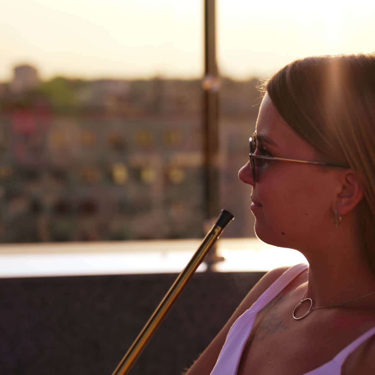 Blonde lady in white dress smoking hookah sitting on the building roof. Woman in sunglasses enjoys shisha looking at the cityscape at sundown