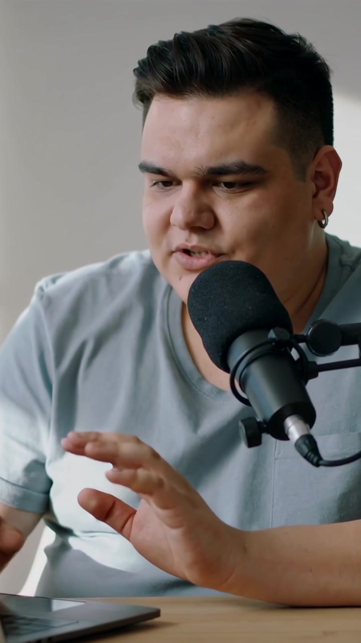 Vertikal video: Vært taster på laptop, iført grå t-shirt, drejer hovedet og taler ind i mikrofon derhjemme.