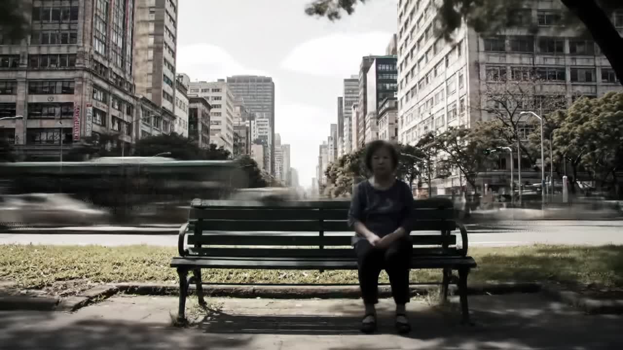 An elderly woman quietly sits on a green bench in a bustling urban center. Skyscrapers surround her while vehicles rush by on the street.