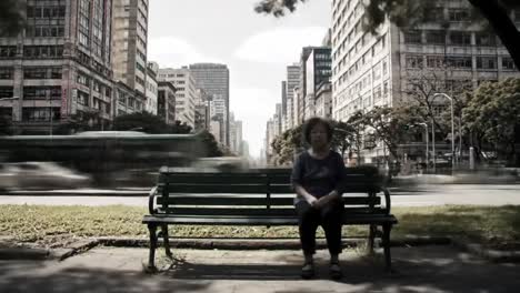 an elderly woman quietly sits on a green bench in a bustling urban center. skyscrapers surround her while vehicles rush by on the street.