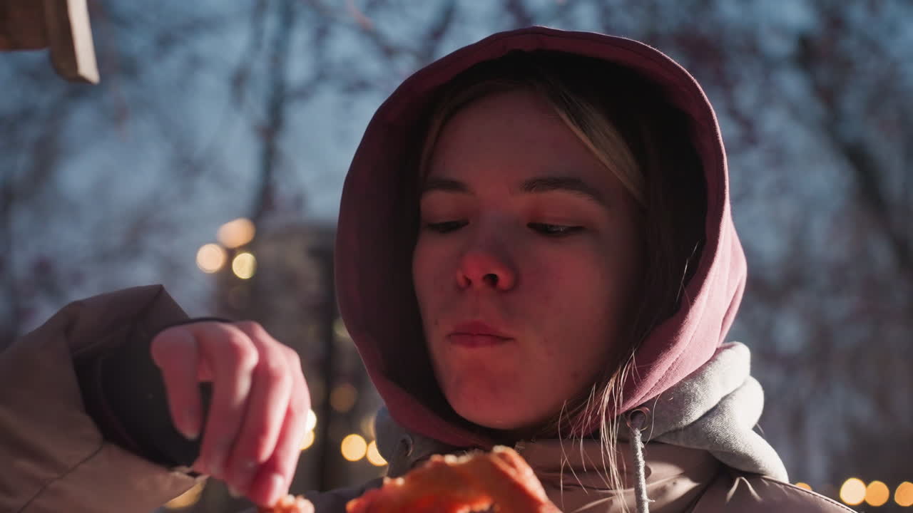 야외에 서 있는 젊은 여성의 클로즈업, 눈이 내린 공원의 새 집에서 새를 위해 음식을 떨어뜨리는 동안 간식을 먹고, 백그라운드에서 빛나는 부드러운 거리 불빛
