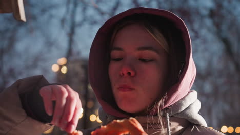 primer plano de una mujer joven de pie al aire libre, comiendo un bocadillo mientras deja caer comida para los pájaros en la casa de pájaros de un parque nevado, luces de la calle suaves brillando en el fondo