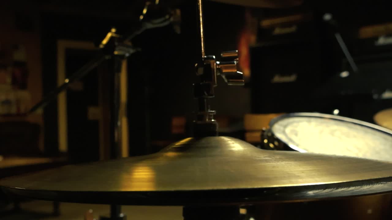 Rotation around an acoustic drum set and cymbals in a dark studio room, where small lights cast a soft glow on the drum kit. The interplay of light and shadow highlights the textures of the cymbals